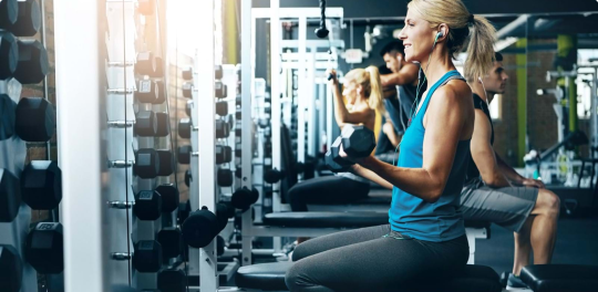 Woman with earbuds in blue tank top sitting on bench doing bicep curls with free weights 