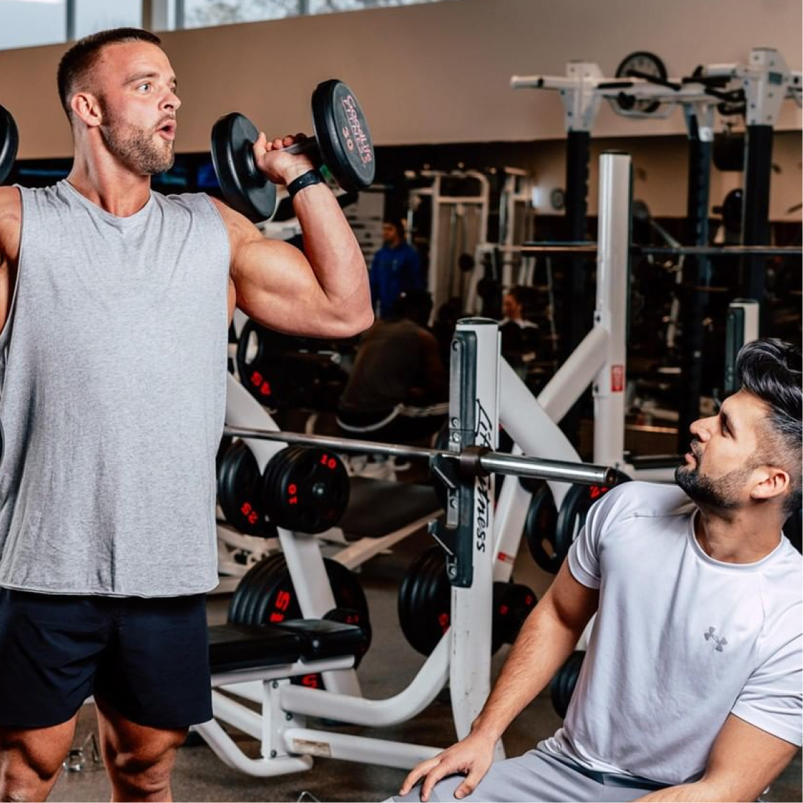 Man in grey shirt does standing shoulder presses with dumbbells while a second man on a bench in a white t-shirt observes his form