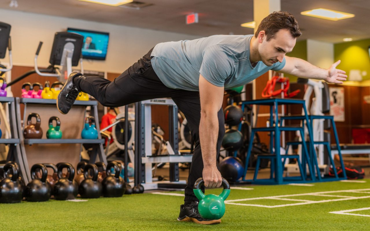 Man holding kettlebell while leaning forward and balancing on one foot on indoor turf
